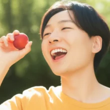 A-smiling-Korean-person-holding-a-ripe-plum-in-a-bright,-sunny-setting,-looking-refreshed-and-happy.